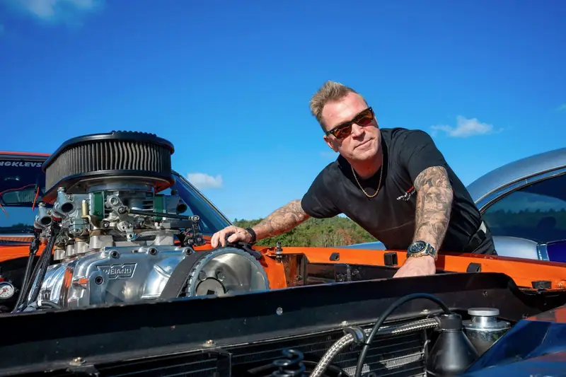 Anders Tångklev i solglasögon lutar sig över det öppna motorrummet på en orange bil med kromad motor, mot en blå himmel.