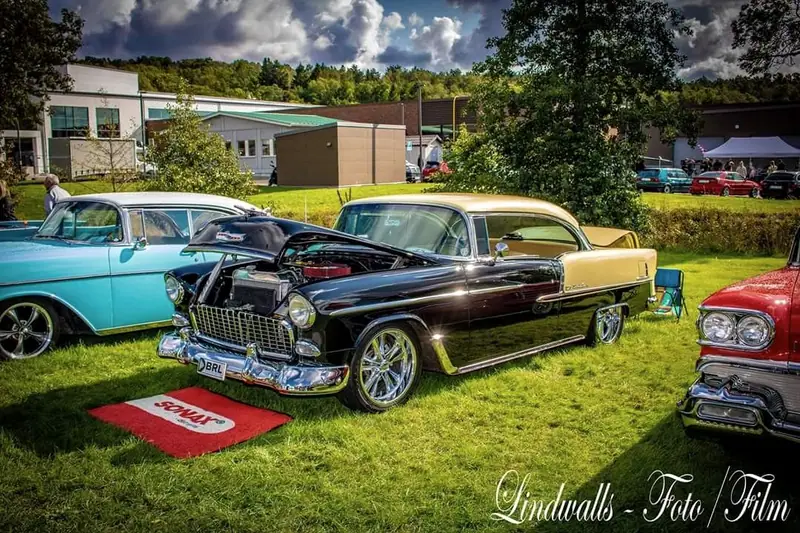 Svart och gul Chevrolet Bel Air med öppen motorhuv på gräset på en bilträff, bland andra amerikanska klassiker.