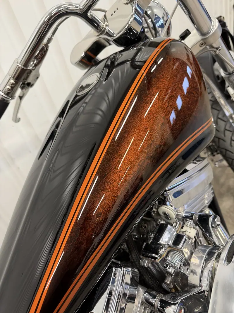 Närbild på motorcykelns tank med orange och svart mönsterlack som glänser i taklamporna.