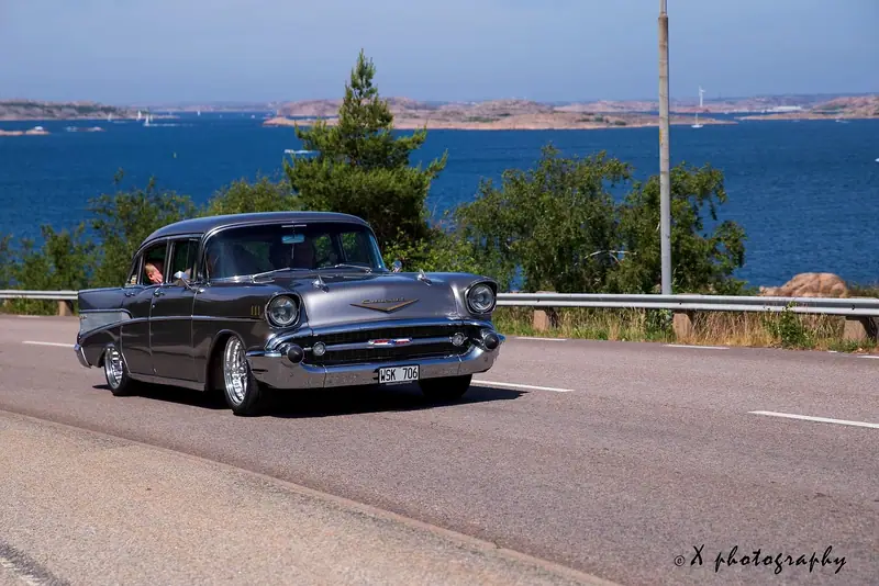 Grå Chevrolet från 1957 kör längs en kustväg med havet och skären i bakgrunden.
