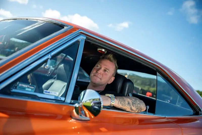 Anders Tångklev sitter bakom ratten i en orange bil och tittar ut genom den öppna sidorutan.
