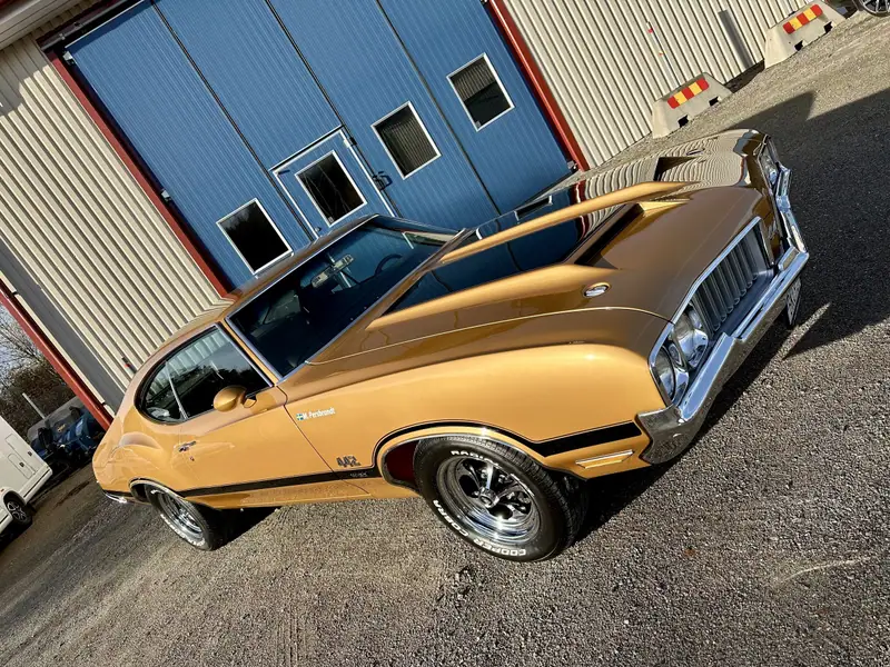 Guldfärgad Oldsmobile 442 snett framifrån framför en byggnad med blå portar.