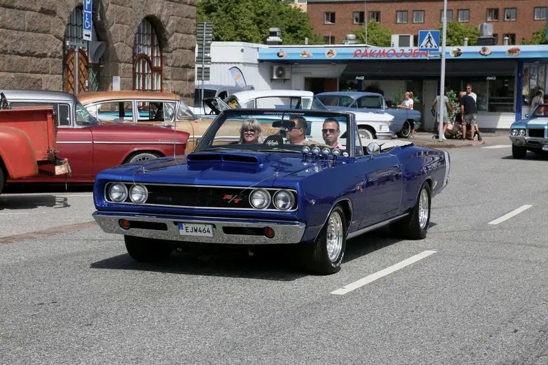 Blå Dodge Coronet R/T cabriolet med tre personer i kör på en gata under en bilträff, med skylten Tångklevs Autoshine på vindrutan.