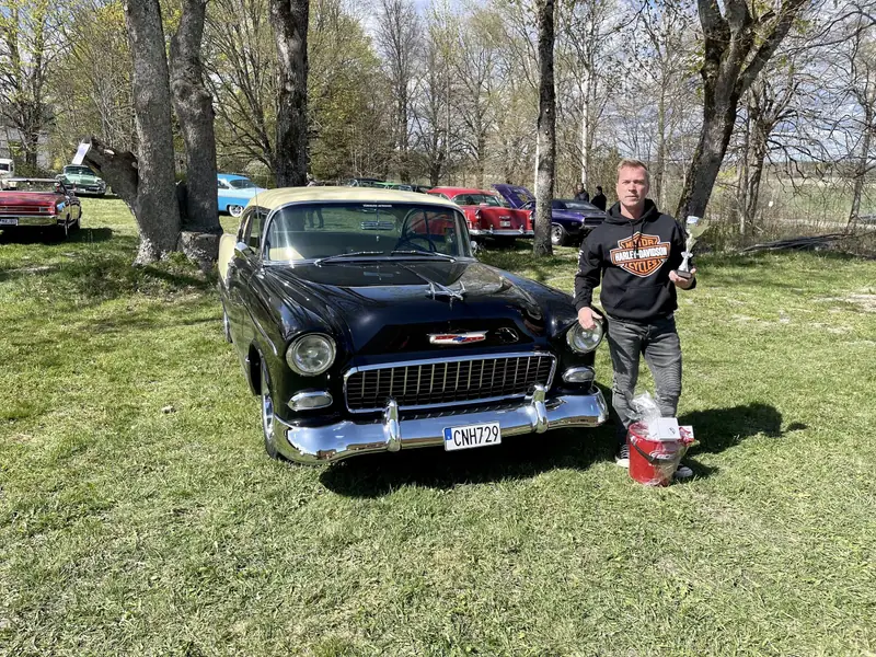 Svart och gul Chevrolet Bel Air från 1955 på en gräsmatta under en bilträff, med en man som håller en pokal bredvid bilen.