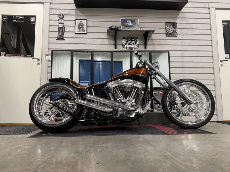 Specialbyggd motorcykel med orange och svart lack, kromade fälgar och brett bakdäck, fotograferad från sidan i ett garage.