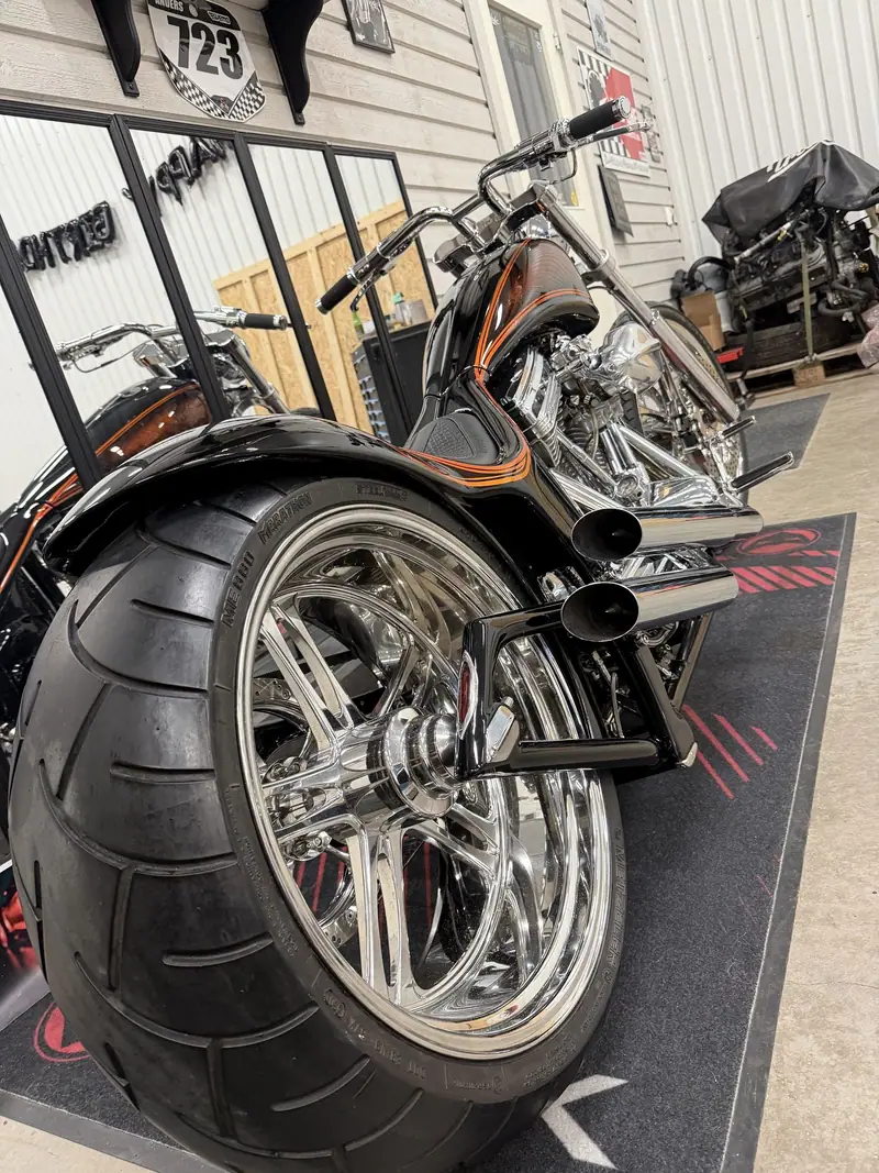 Motorcykelns breda bakhjul med kromad fälg och blanka ljuddämpare, fotograferat snett bakifrån.