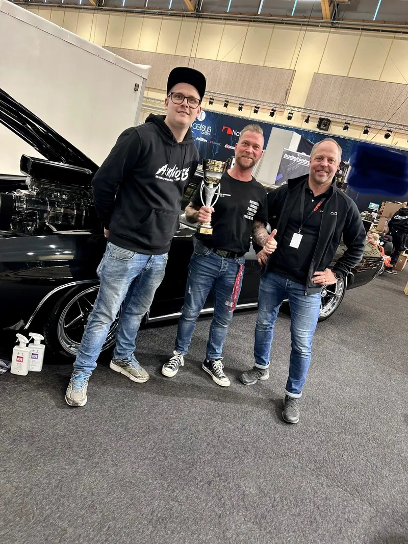 Tre män framför en svart bil med öppen motorhuv på en bilmässa. Mannen i mitten håller en pokal.