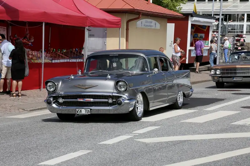 Grå Chevrolet från 1957 på en gata under en bilträff, med marknadsstånd i bakgrunden.