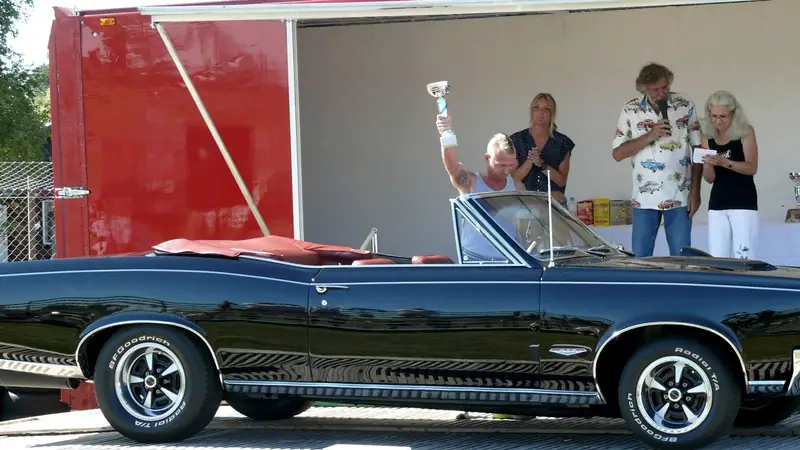En man håller upp en pokal i en svart cabriolet framför scenen under en prisutdelning på en bilträff.