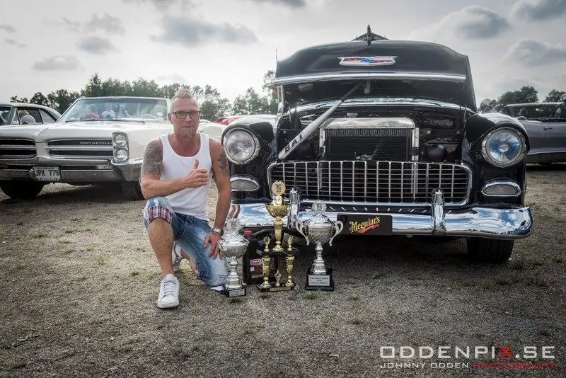 Anders Tångklev på knä med pokaler framför en svart och gul Chevrolet Bel Air med öppen motorhuv på en bilträff.