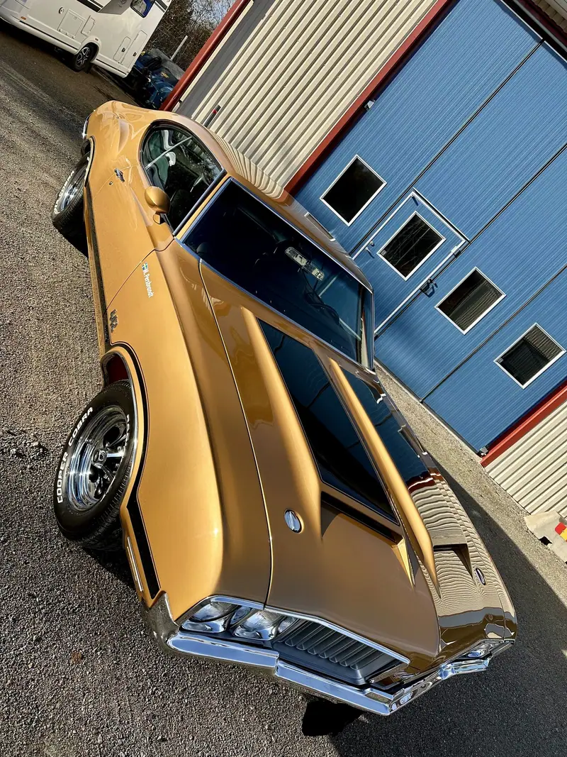 Guldfärgad Oldsmobile 442 fotograferad snett ovanifrån framför verkstadens blå portar.