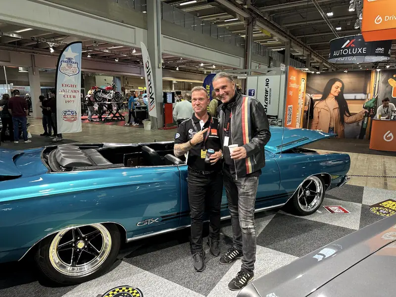 Två män står framför den blå Plymouth GTX-cabrioleten i montern på Oslo Motor Show 2025.