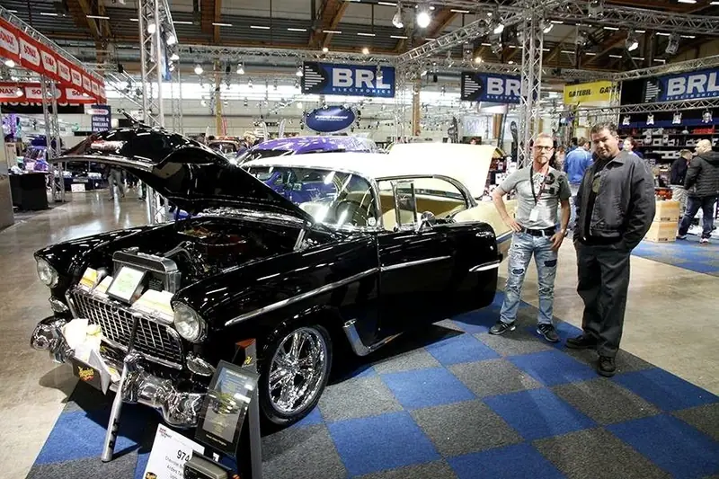 Två män står bredvid en svart Chevrolet Bel Air med öppen motorhuv i en monter på en bilmässa.