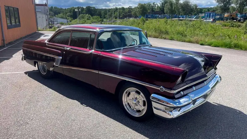Vinröd amerikansk 50-talsbil med blank lack och kromad stötfångare på en asfaltsplan i solsken.