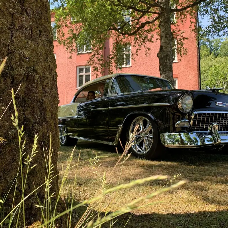 Svart Chevrolet Bel Air från 1955 med blank lack och krom, parkerad i gräset under ett träd framför ett rött tegelhus.