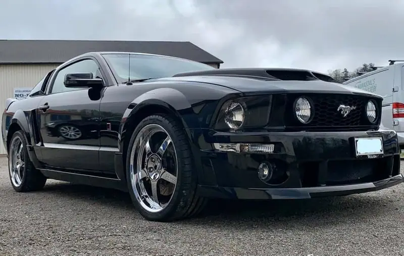 Svart Ford Mustang med blank lack på en grusplan under en grå himmel.