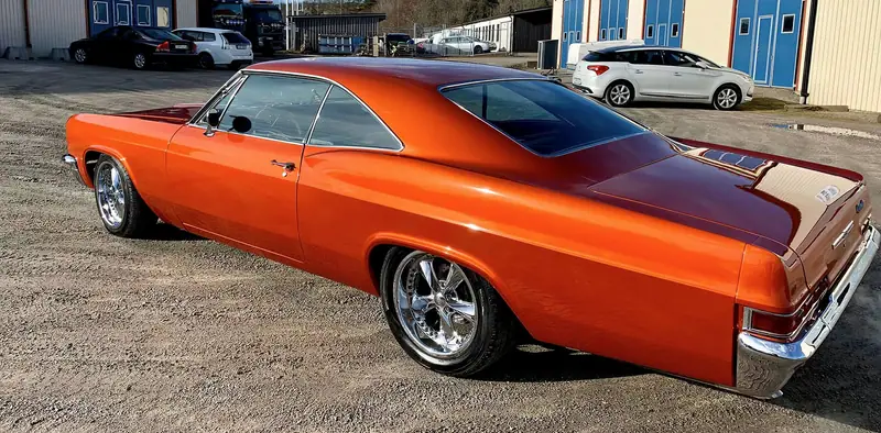 Orange amerikansk 60-talsbil med kromade fälgar, fotograferad snett bakifrån på grusplanen vid verkstaden.