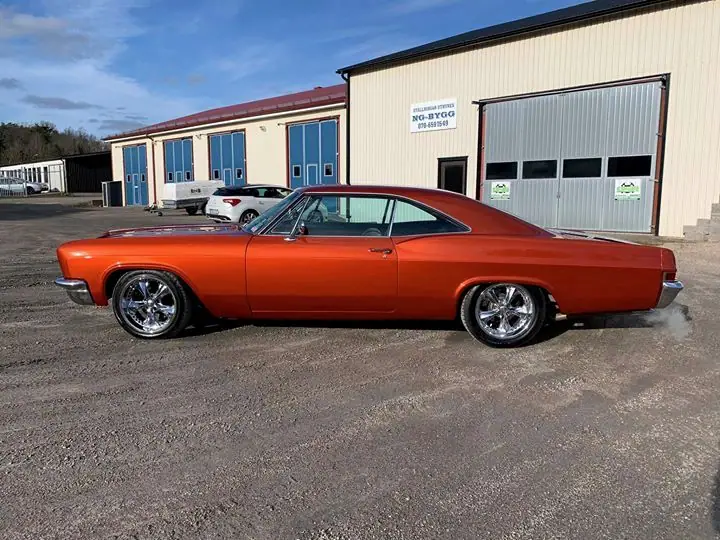 Orange amerikansk 60-talsbil med kromade fälgar, fotograferad från sidan framför verkstaden.