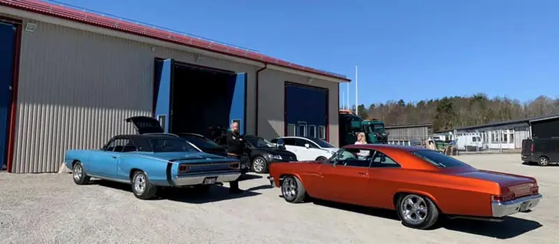 En ljusblå och en orange amerikansk bil parkerade utanför verkstadens öppna port en solig dag.