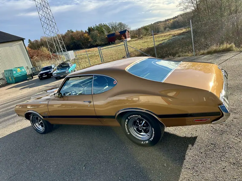 Guldfärgad Oldsmobile 442 med svarta sidostripes på en grusplan i kvällssol.