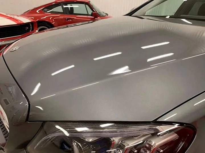 Närbild på en grå motorhuv där verkstadens lampor speglas i lacken, med en röd sportbil i bakgrunden.
