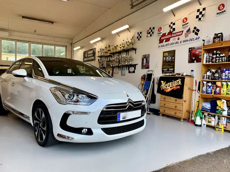 Vit Citroën DS5 med svart tak i verkstaden, framför hyllor med bilvårdsprodukter och rutiga flaggor.