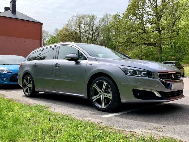 Grå Peugeot 508 SW kombi parkerad vid en gräsmatta med grönska i bakgrunden.